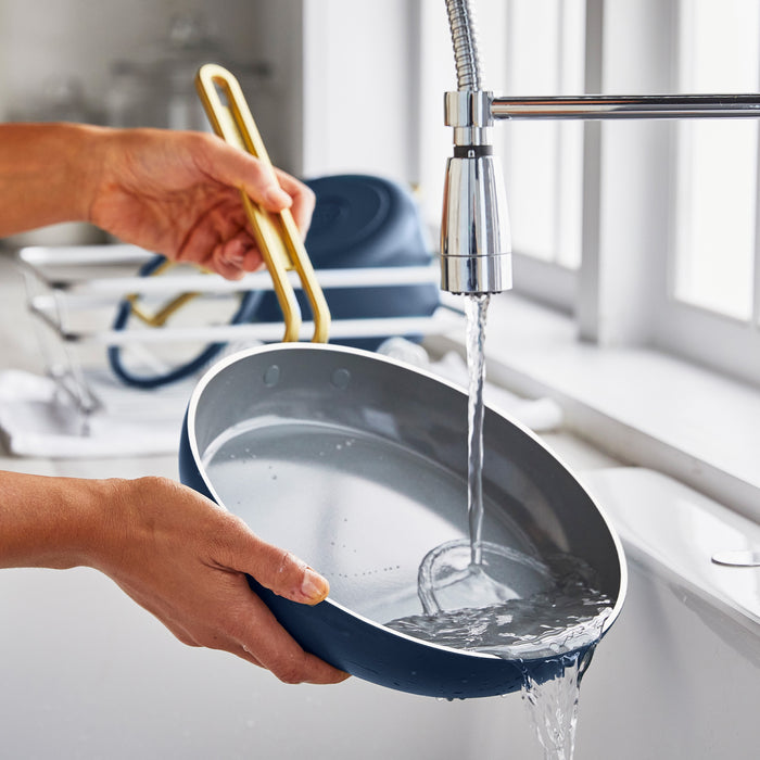 Person rinsing a Marino Blue ceramic nonstick 8-inch frypan under running water in a kitchen sink by a window.