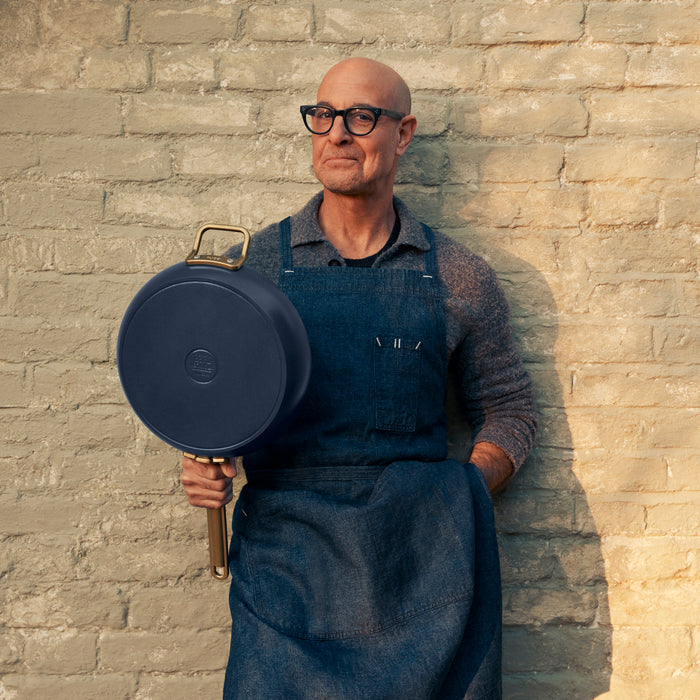 Man in glasses and blue apron holding a Marino Blue Stanley Tucci ceramic nonstick 8 frypan in front of a brick wall.