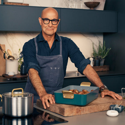 Man wearing glasses and an apron holding lasagna in a white Stanley Tucci™ ceramic lasagna pan in a modern kitchen.