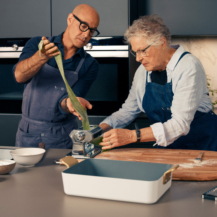 Two people roll green dough with a pasta maker next to a teal Stanley Tucci Lasagna Pan.
