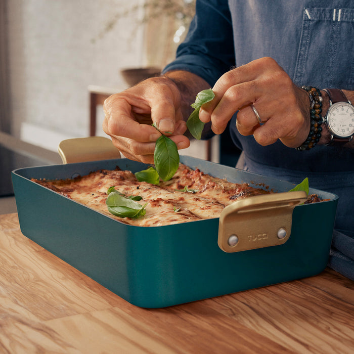 Person adding basil to lasagna in a teal ceramic nonstick lasagna pan on a wooden table.