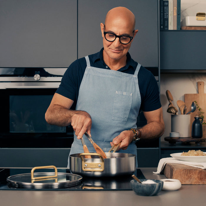 Man wearing glasses and an apron uses wooden utensils with a stainless steel 4-quart sauté pan on the stove.