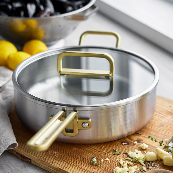 Stainless steel sauté pan with lid on a board, next to chopped garlic and fresh herbs. 4-quart size.
