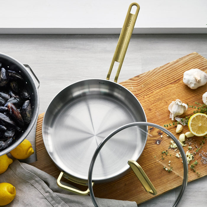 Stainless steel 4-quart sauté pan with gold handle and lid, resting on a board with mussels and fresh herbs.