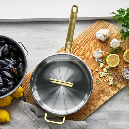 Stainless steel sauté pan with lid on a wooden board, with lemons and herbs around it.