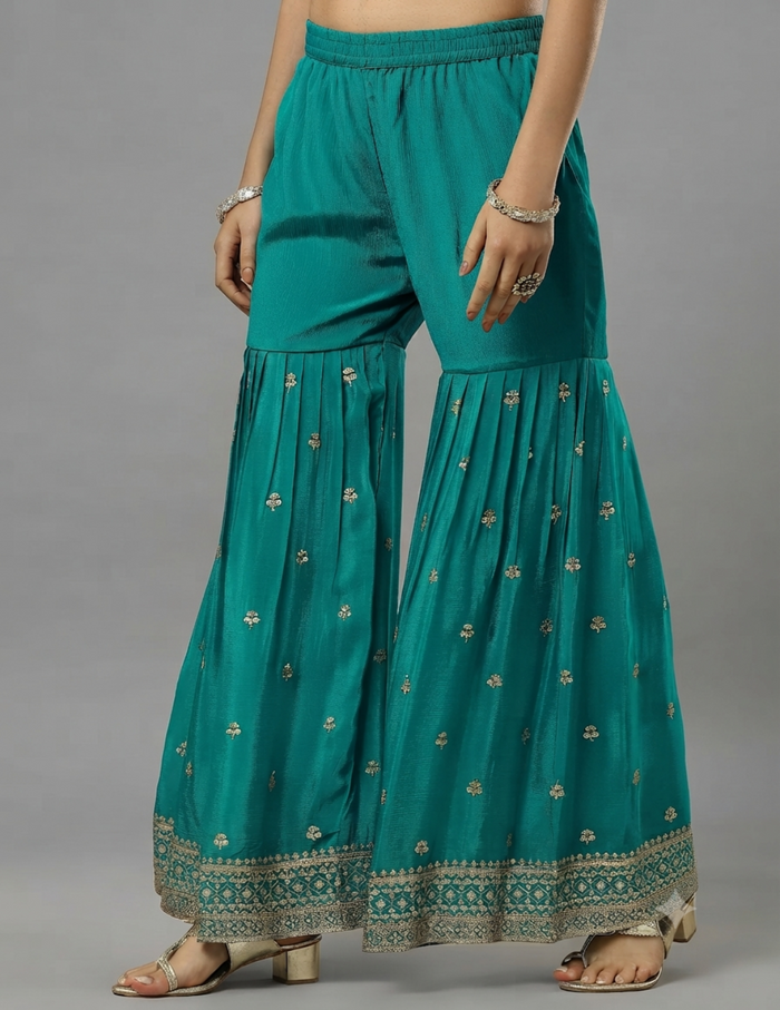 A model is wearing teal green wide-leg palazzo pants with small gold floral embroidery and a decorative gold border at the hem, paired with gold sandals and jewelry, set against a solid gray studio background.
