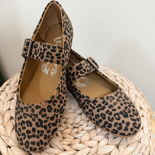 A pair of women's Mary Jane flats with a tan and black leopard print, resting on a woven straw stool against a white wall.
